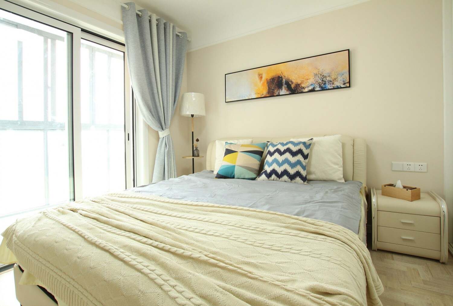 主卧采用浅色系的床上四件套、壁纸和淡蓝色的窗帘，简单干净又温馨。床上的几何抱枕和抽象挂画色彩艳丽。